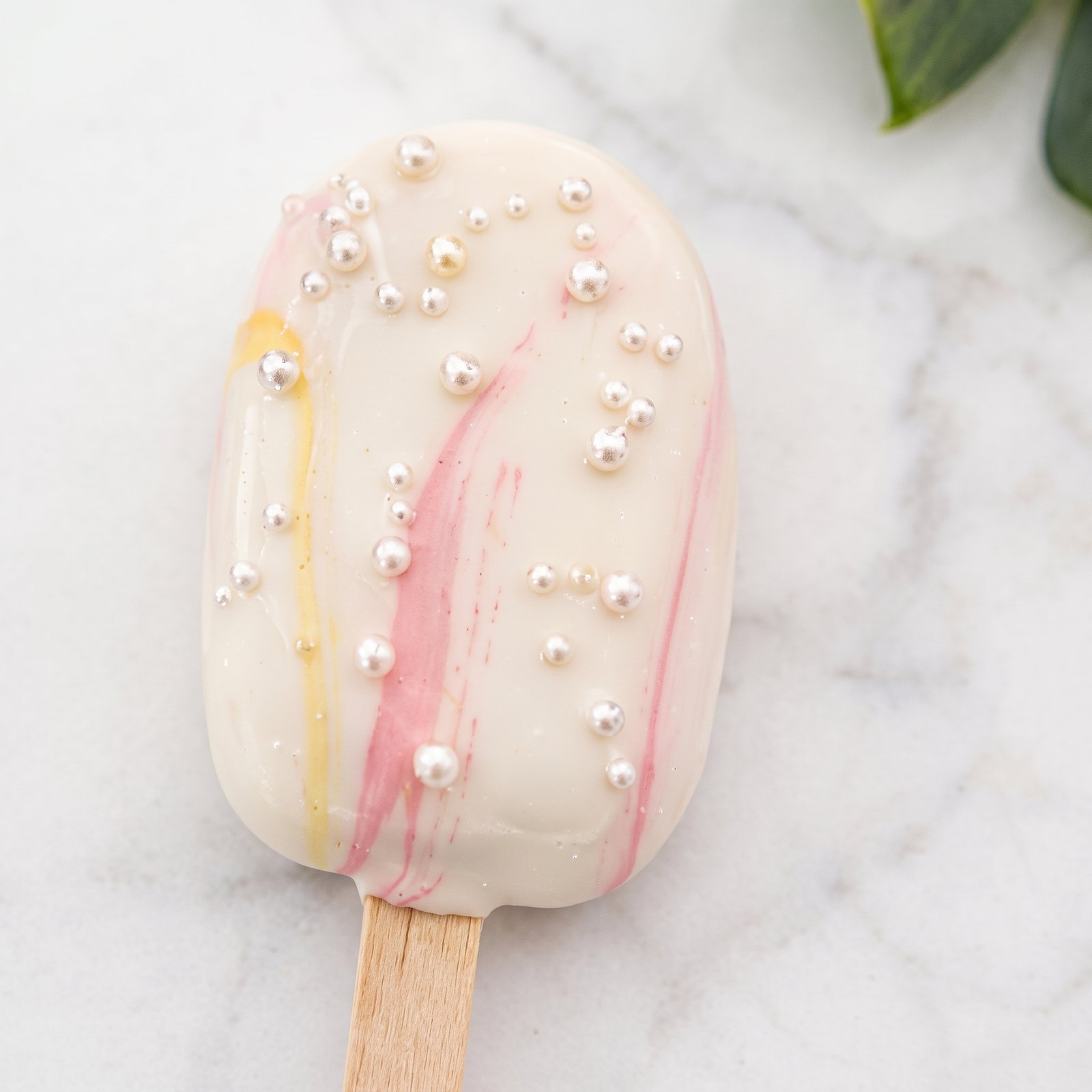 A white cakesicle with pink drizzle and pearls on marble